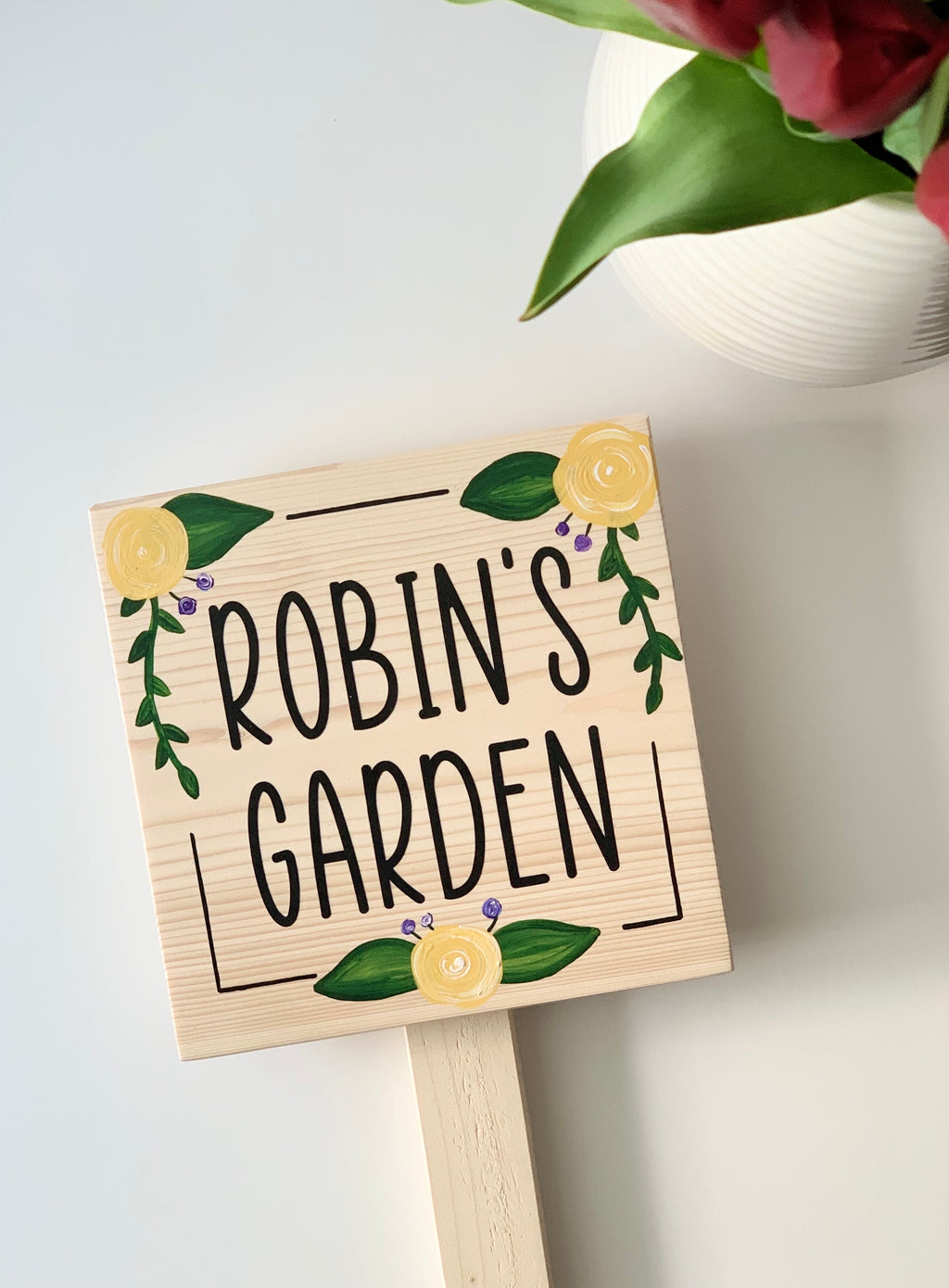 Small square natural wooden garden sign with black letters and yellow floral illustrations on stake white background