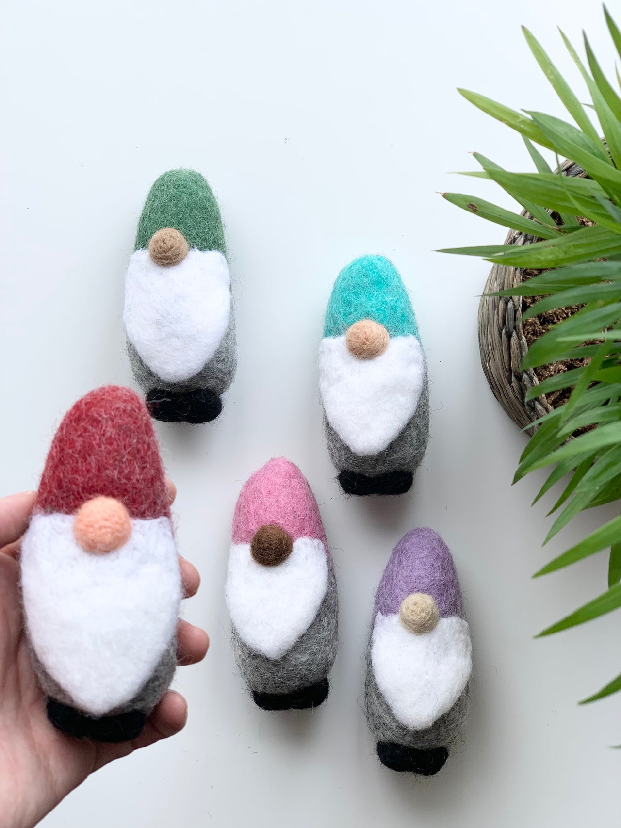 Handmade felt gnome with red hat, white beard, gray body, and black shoes held near four other colorful felt gnomes on white background