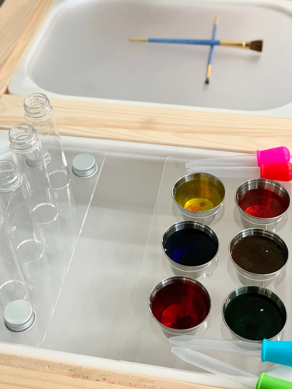 Clear acrylic test tube Flisat insert with metal lids, metal cups filled with colorful liquids, and droppers on wooden table