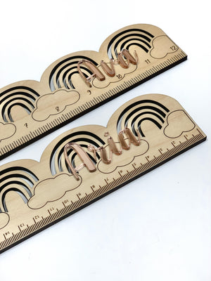 Natural wooden personalized rainbow ruler with engraved black cloud and rainbow design and cursive rose gold name on white background