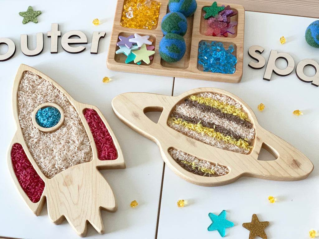 Natural wooden rocket and Saturn planet sensory trays filled with colorful textured materials on white surface with star decorations