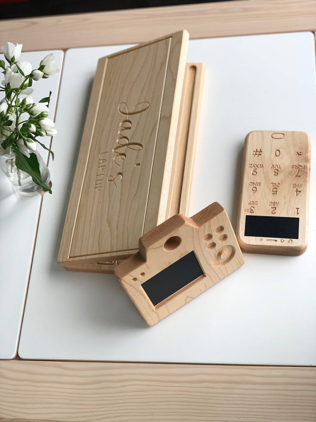 Natural wooden toy camera and phone with engraved details and black screens next to engraved wooden box on white surface
