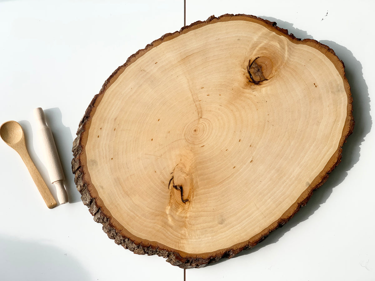 Natural live edge wooden tray with smooth clear coat and bark edges beside small wooden spoon and rolling pin on white surface