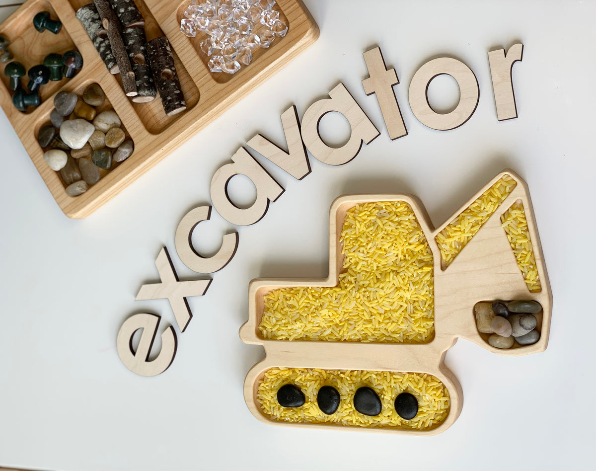 Wooden excavator-shaped sensory tray filled with yellow rice and small rocks near natural elements on white surface