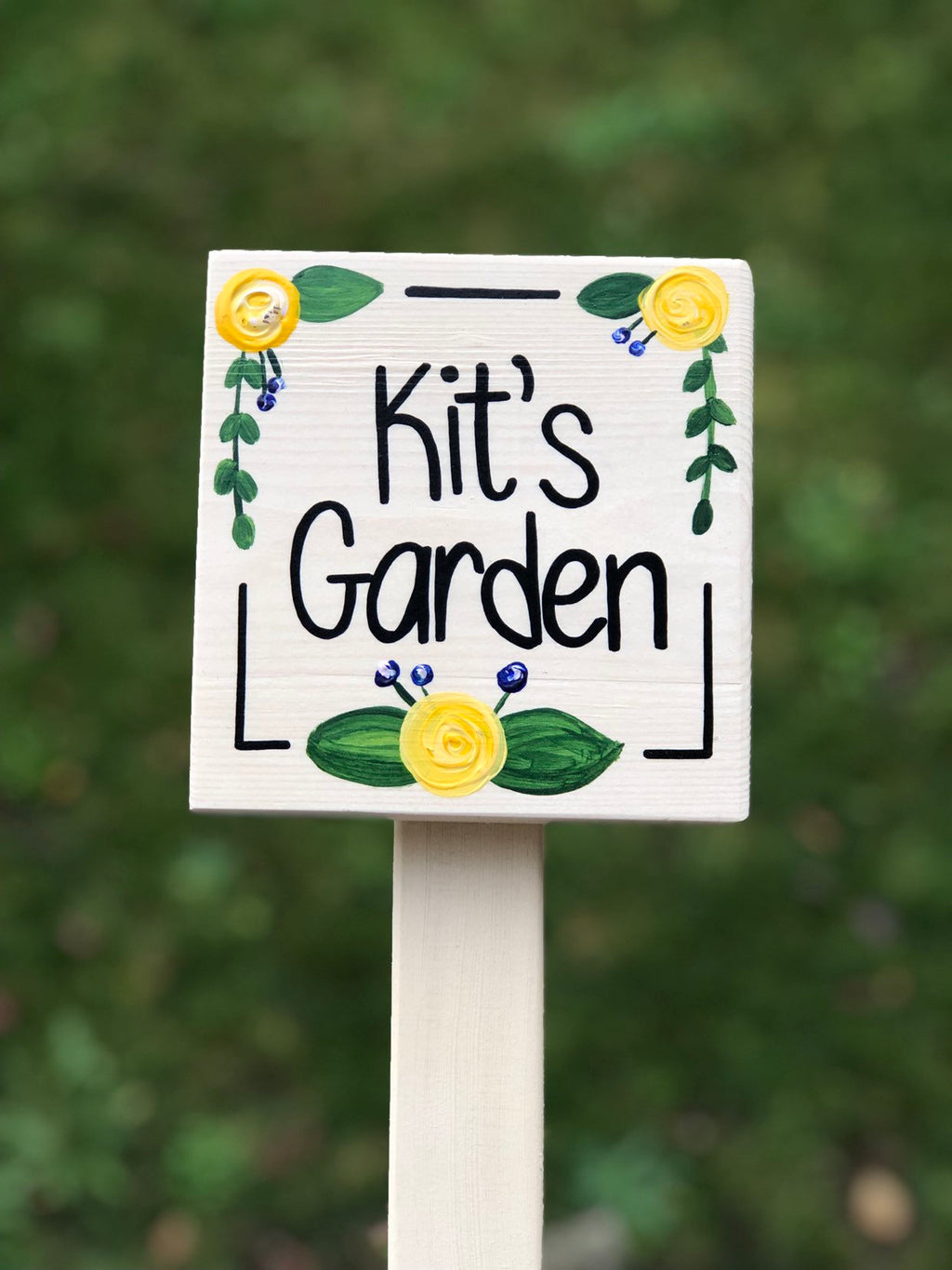 Small square wooden garden sign painted white with black lettering and yellow floral accents on stake background blurred green grass