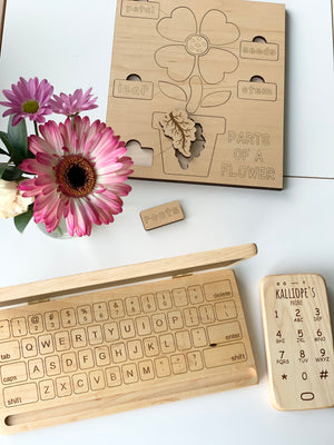 Natural wooden educational puzzle labeled parts of a flower with engraved flower pieces and wooden toy phone and keyboard nearby