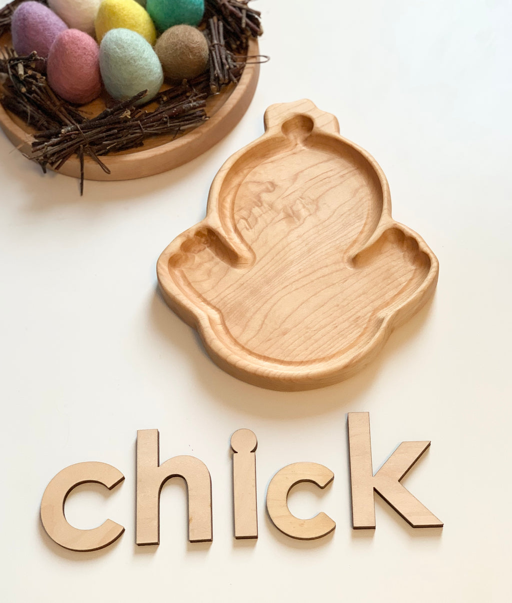 Wooden chick-shaped sensory tray with natural wood letter tiles spelling chick and felt eggs in a small twig nest