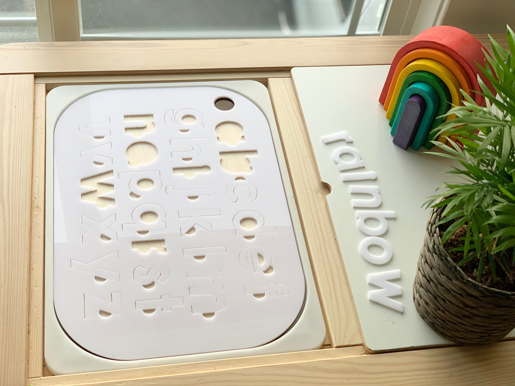 Lowercase alphabet puzzle Flisat tabletop insert with white acrylic letters on maple plywood base next to wooden rainbow toy and plant