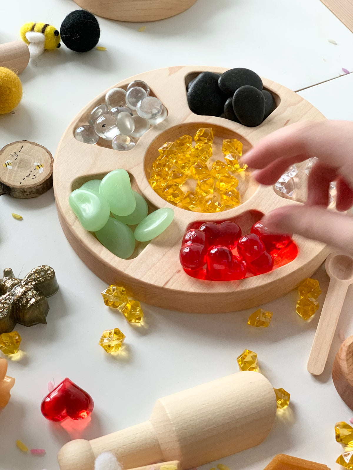 Round wooden tinker tray with compartments holding colorful sensory stones and small play objects on white surface