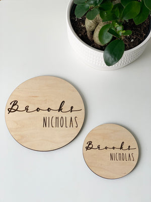 Two personalized round wooden baby name announcement discs with engraved Brooks Nicholas on white surface near potted plant