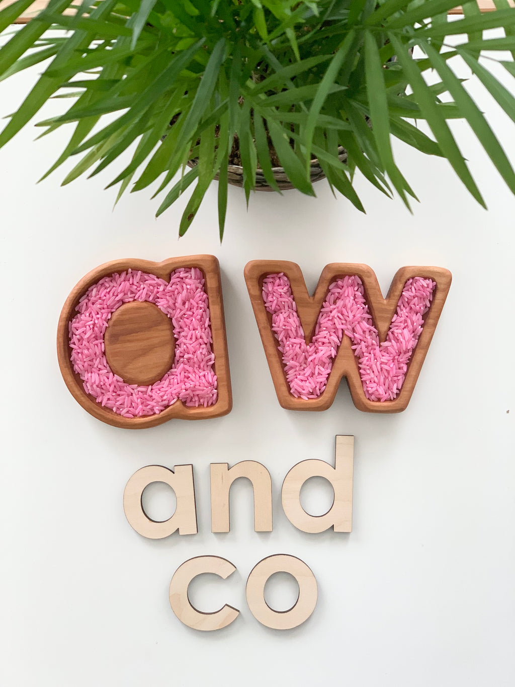 Small wooden alphabet trays filled with pink rice forming letters A and W with wooden letters spelling and on white surface