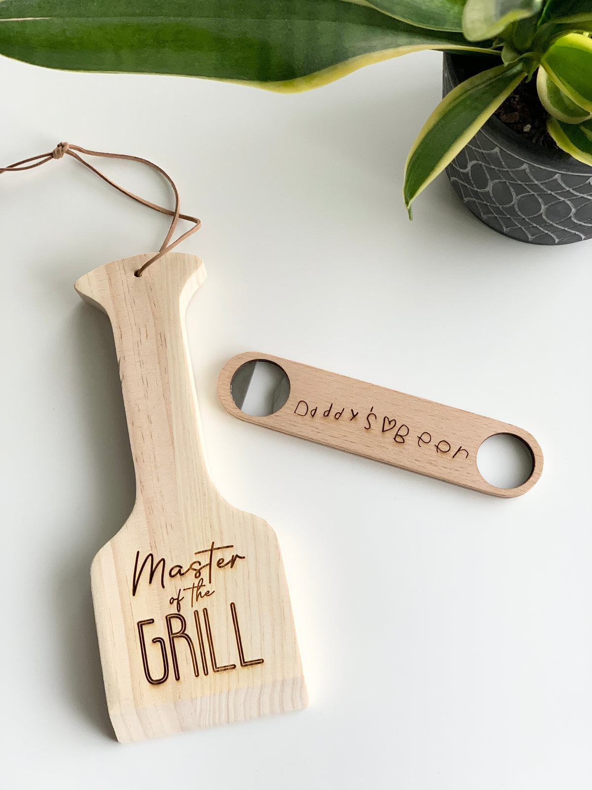 Custom wooden barbecue scraper with "Master of the Grill" engraving and personalized handwritten wooden bottle opener on white surface