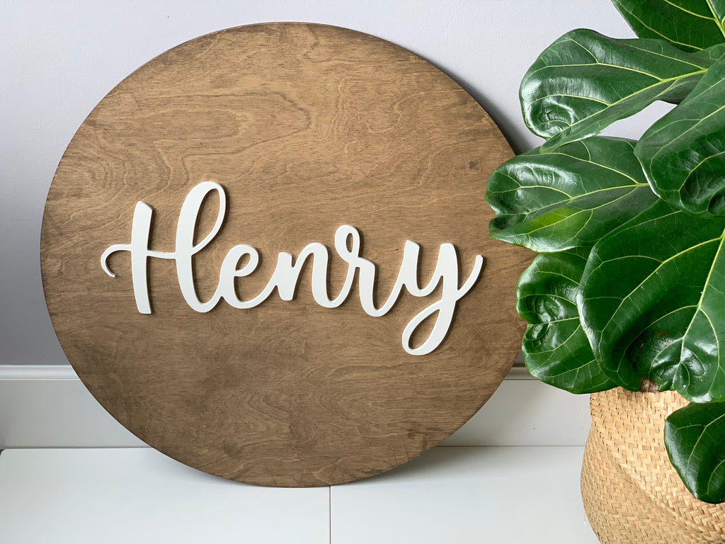 Large round wooden name sign stained brown with white cursive letters spelling Henry near green potted plant