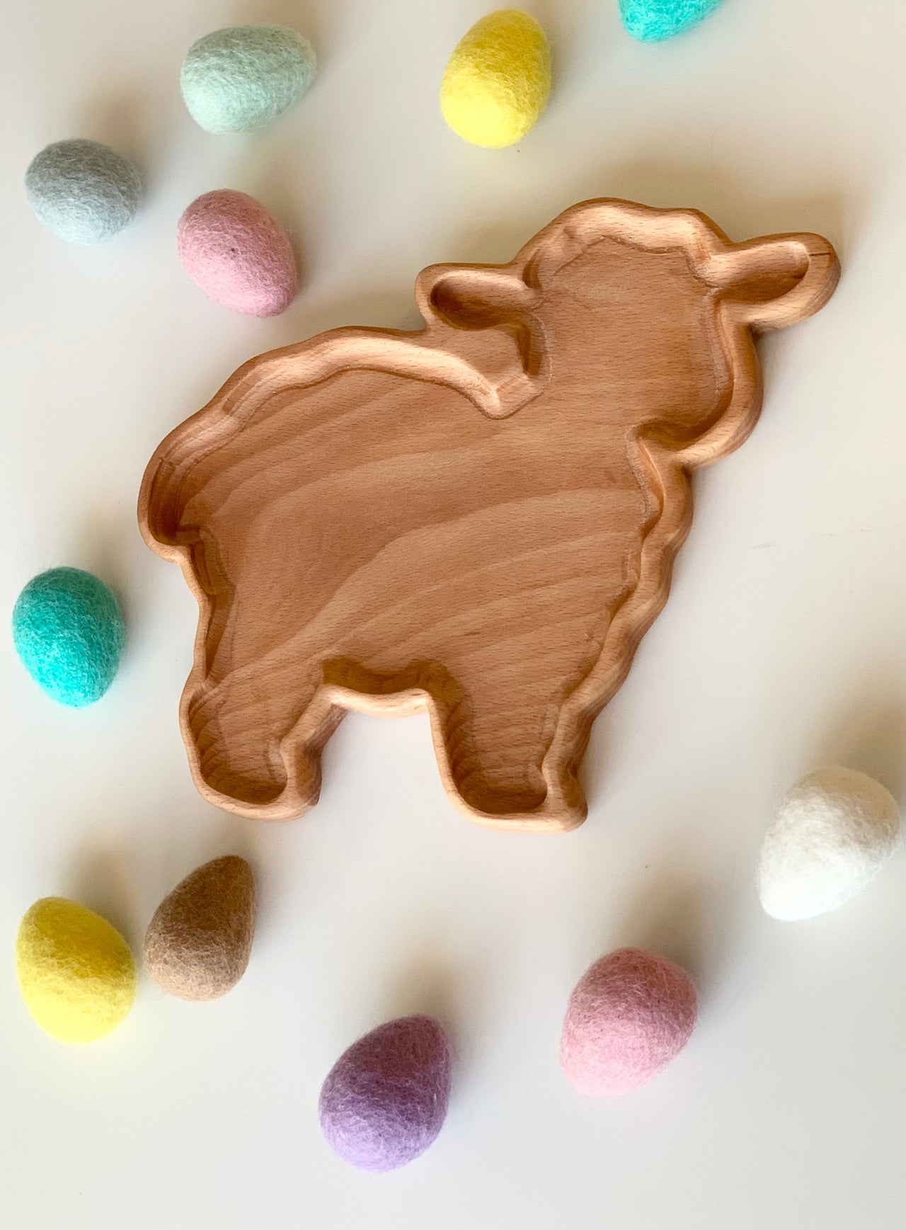 Wooden lamb-shaped sensory tray surrounded by pastel-colored felt eggs on white surface