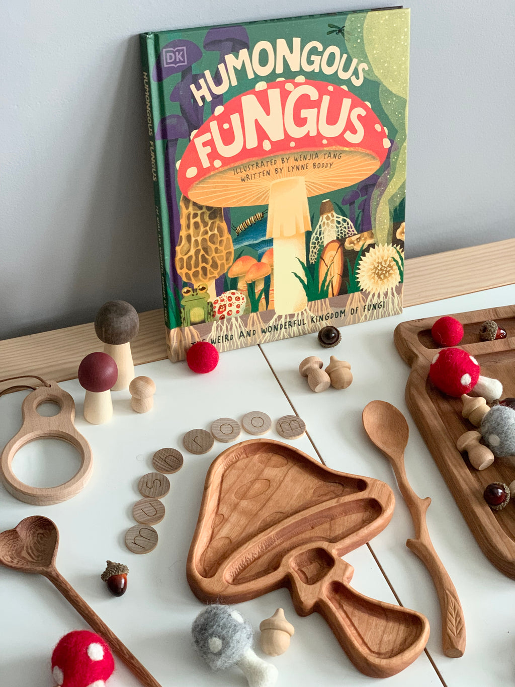 Wooden mushroom-shaped sensory tray with natural wood spoons, felt acorns, alphabet blocks, and a colorful fungi-themed children's book on wooden table
