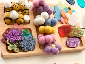 Natural rectangular wooden tinker tray with compartments holding colorful glitter flowers, felt bees, pom poms, and wooden shapes