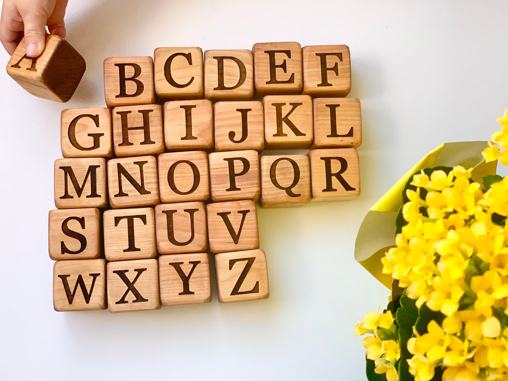 Natural 1.5-inch wooden alphabet blocks with engraved capital letters arranged on white surface near yellow flowers