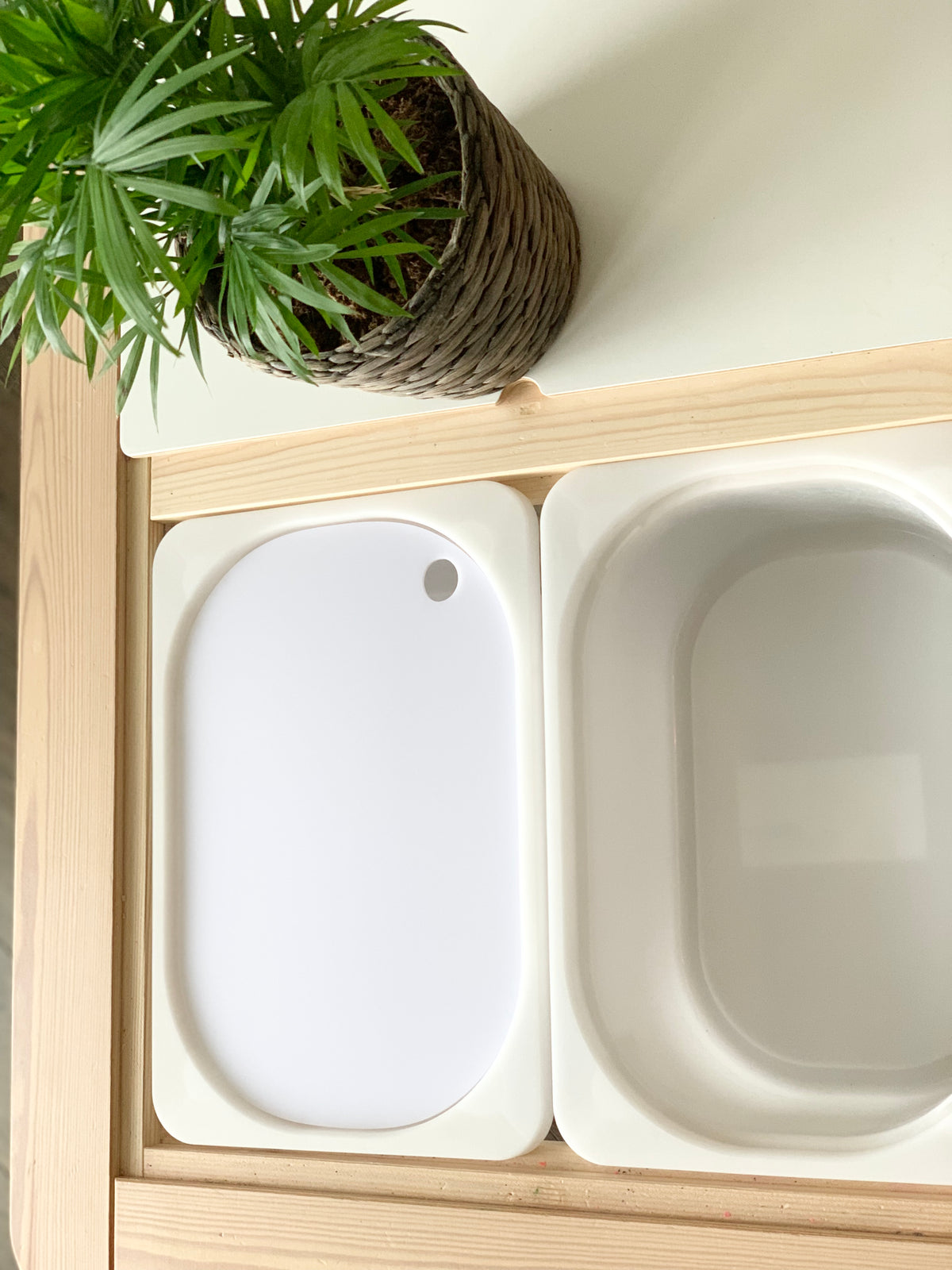 White acrylic Flisat table bin lid with rounded corners next to natural wood frame and green potted plant
