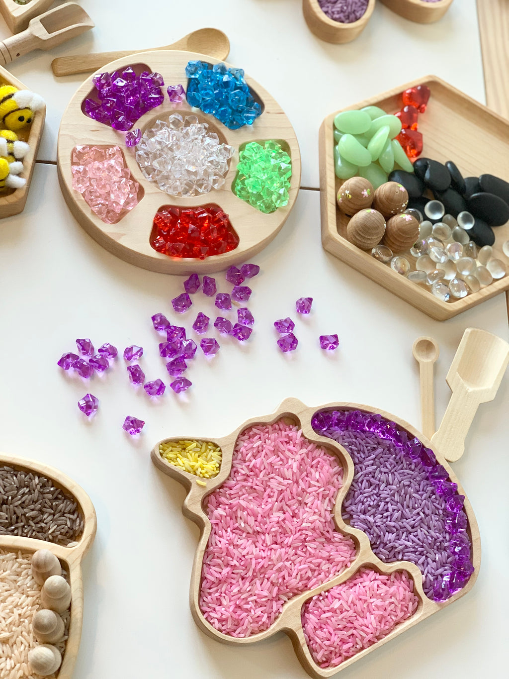 Wooden unicorn-shaped sensory tray filled with colored rice and purple gems, surrounded by wooden tools and colorful beads on white surface