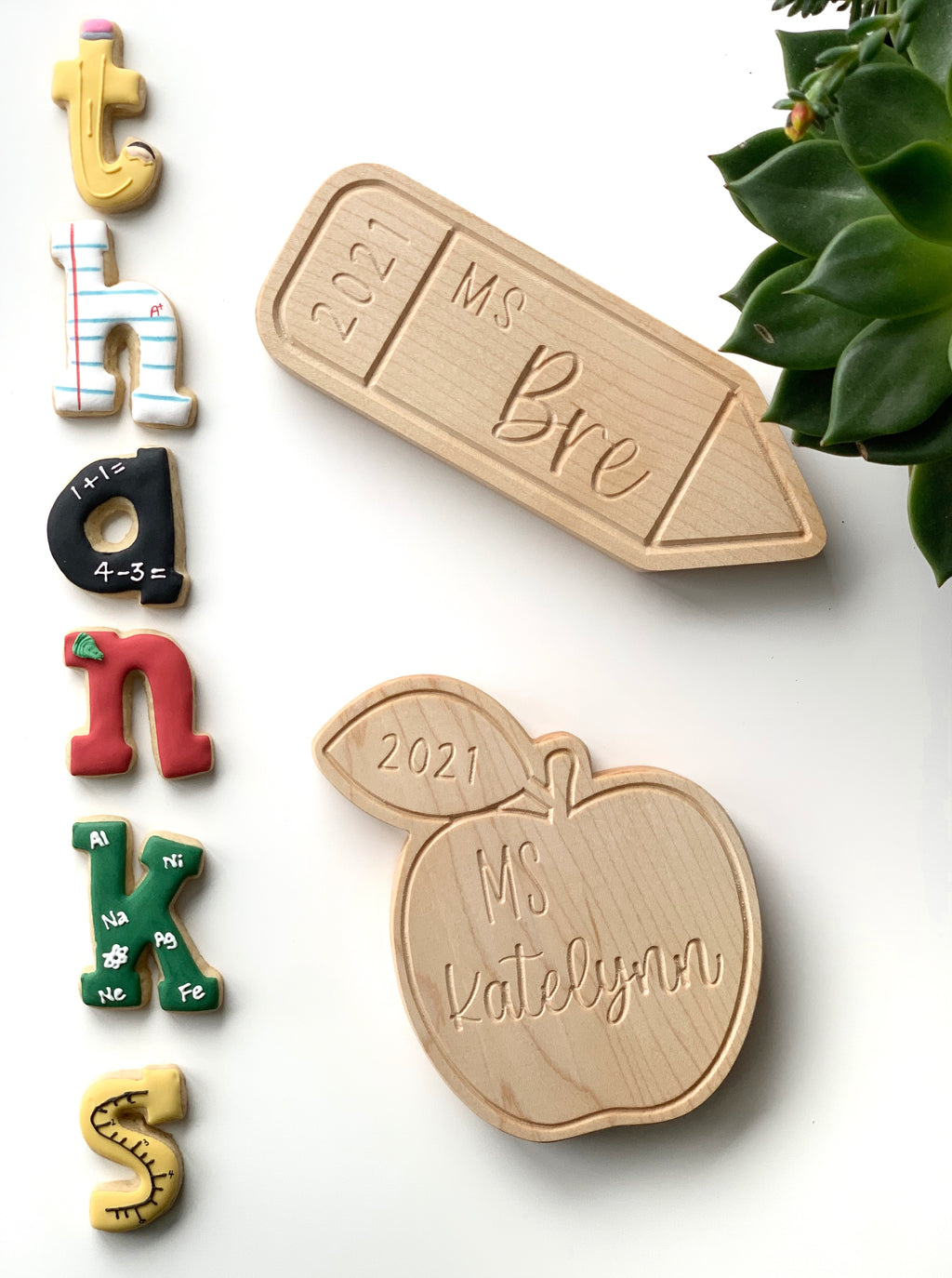 Two personalized wooden teacher name plates shaped like a pencil and an apple on white background with colorful alphabet magnets and green plant