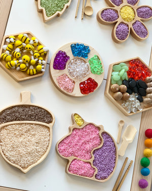 Wooden sensory trays shaped like acorn, unicorn, flower, hexagons filled with colorful rice, beads, and felt bees on white table