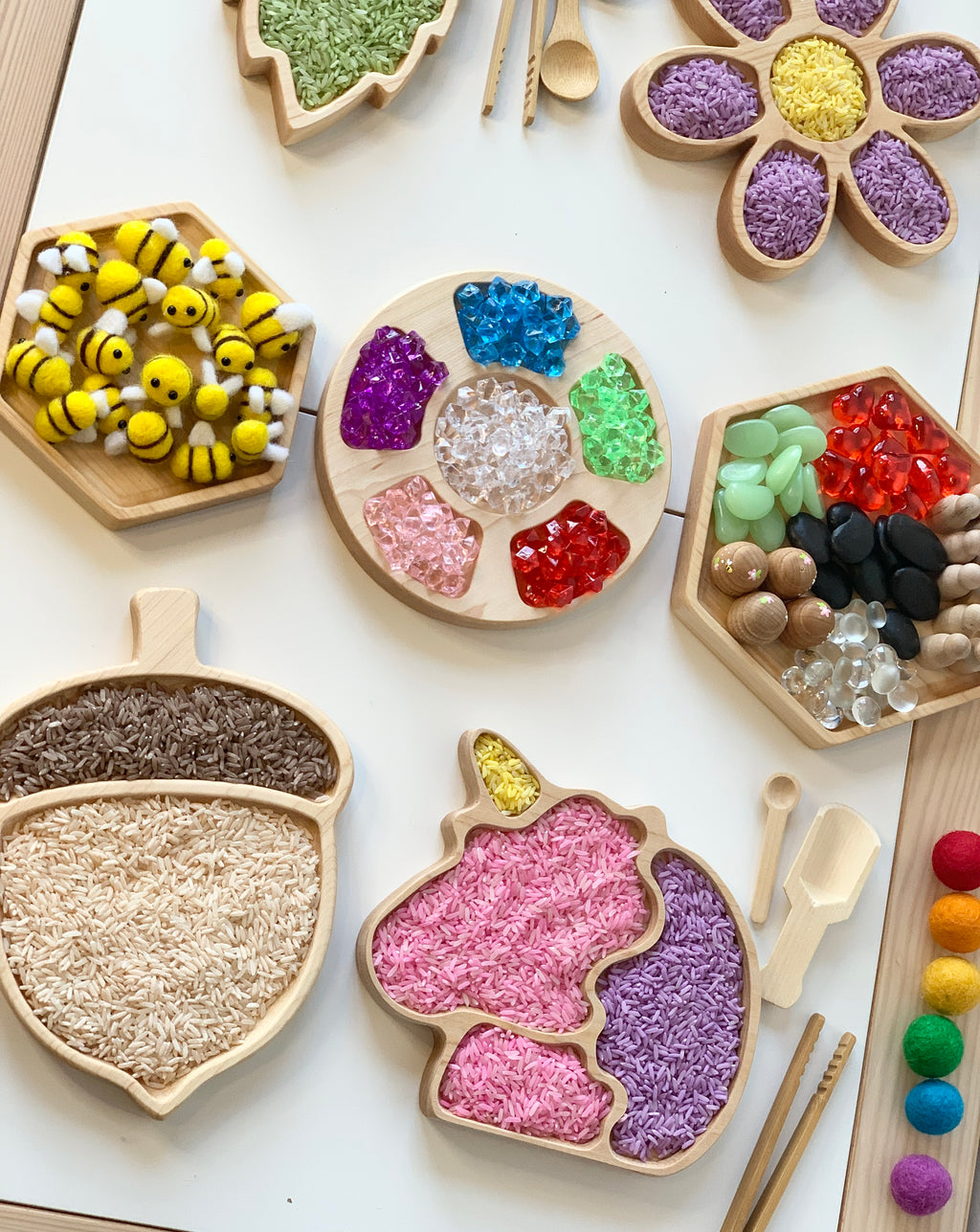 Wooden sensory trays shaped like acorn, unicorn, flower, hexagons filled with colorful rice, beads, and felt bees on white table