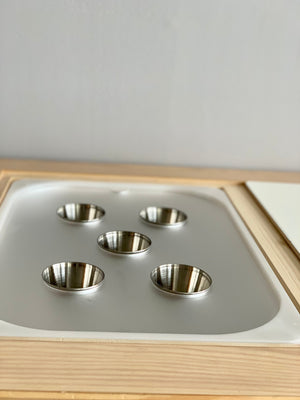 Wooden sensory tray with white acrylic insert holding five small shiny metal cups on light wood surface