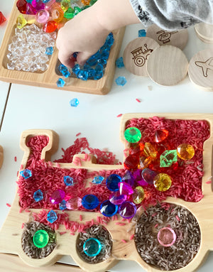 Wooden train-shaped sensory tray filled with colored rice and assorted colorful plastic gems on white surface