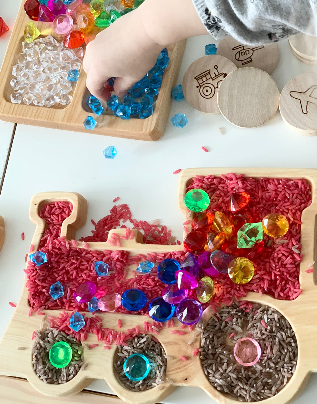Wooden train-shaped sensory tray filled with colored rice and assorted colorful plastic gems on white surface