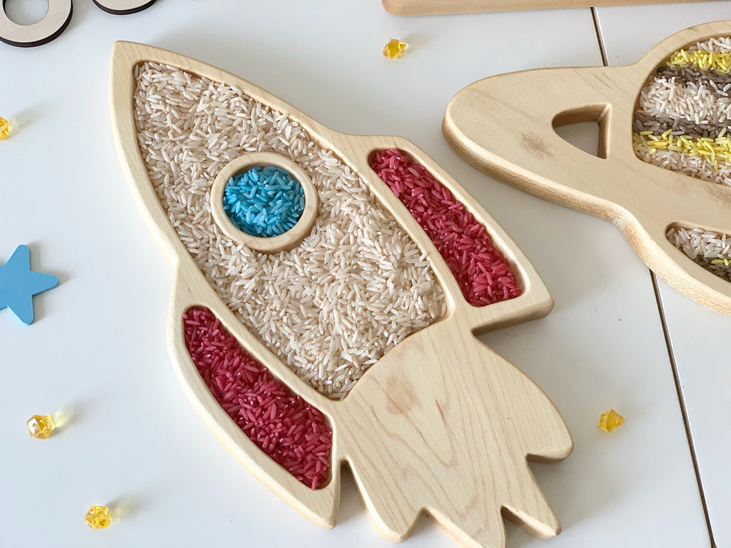 Natural wooden rocket ship sensory tray filled with colored rice on white surface with blue star toy nearby
