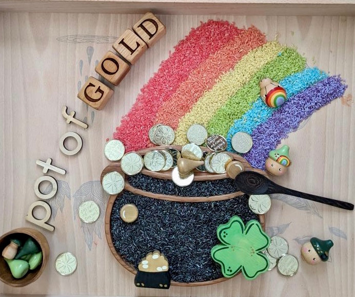 Wooden sensory tray with rainbow colored rice, black rice cauldron plate, wooden coins and St Patrick’s Day themed figures