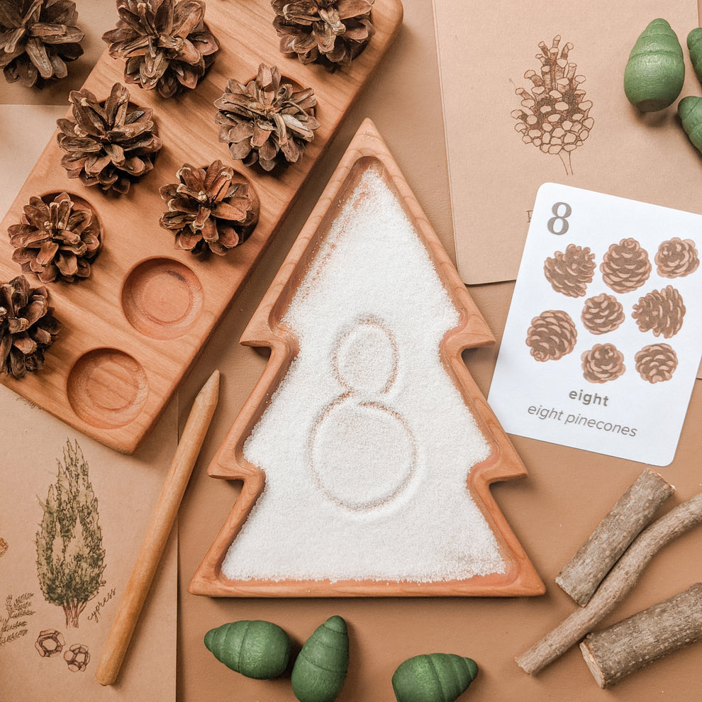 Natural wooden tree-shaped sensory tray filled with salt and number eight drawn inside with pinecones and nature cards nearby