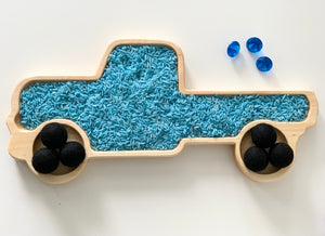 Natural wooden truck-shaped sensory tray filled with blue rice and black felt balls as wheels on white surface