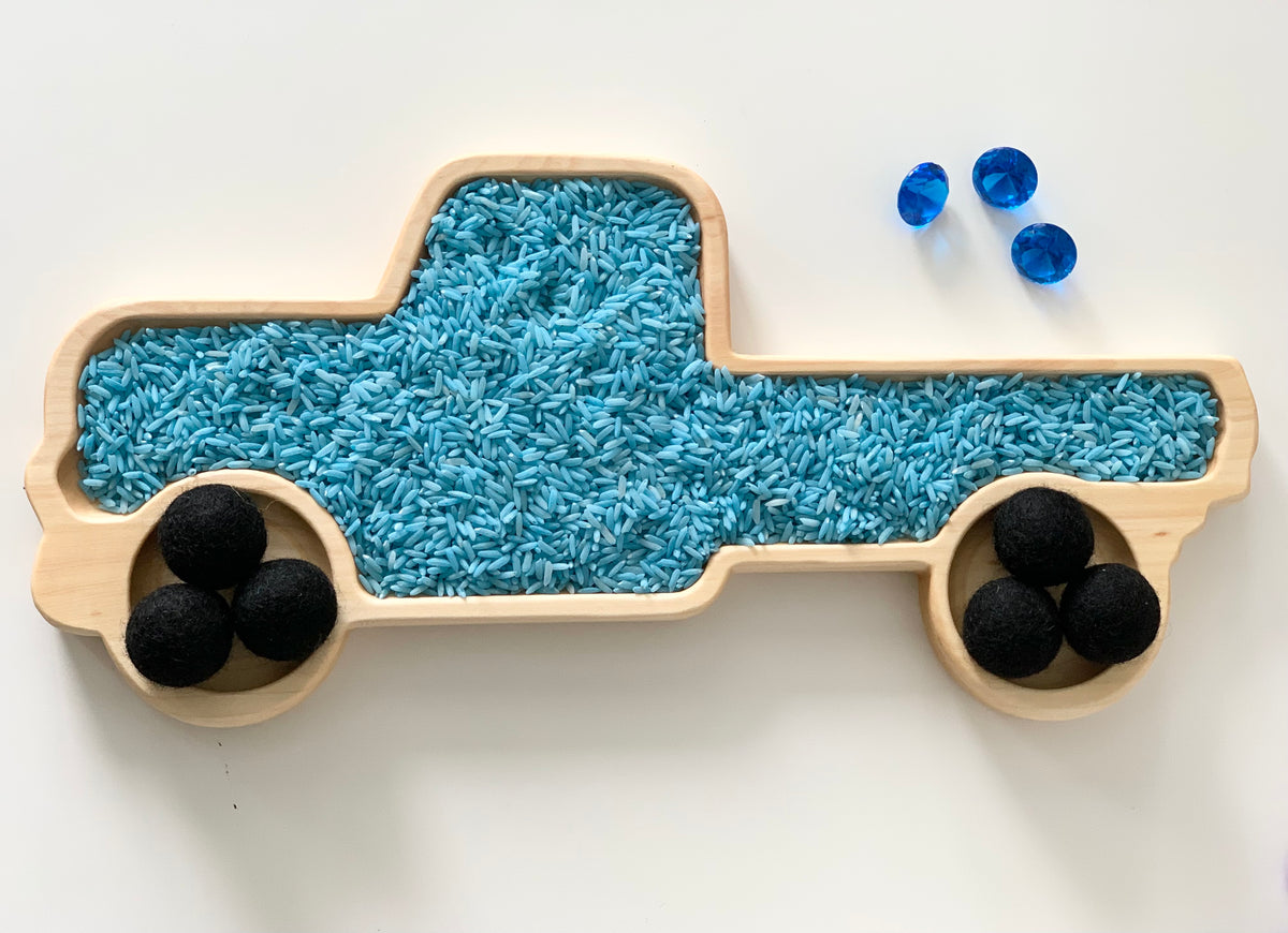 Natural wooden truck-shaped sensory tray filled with blue rice and black felt balls as wheels on white surface