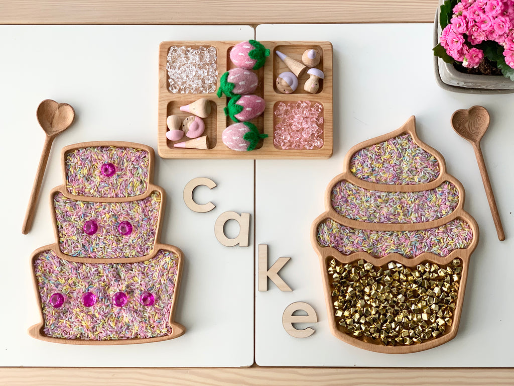 Wooden cake-shaped and cupcake-shaped sensory trays with textured materials and small wooden spoons on white surface