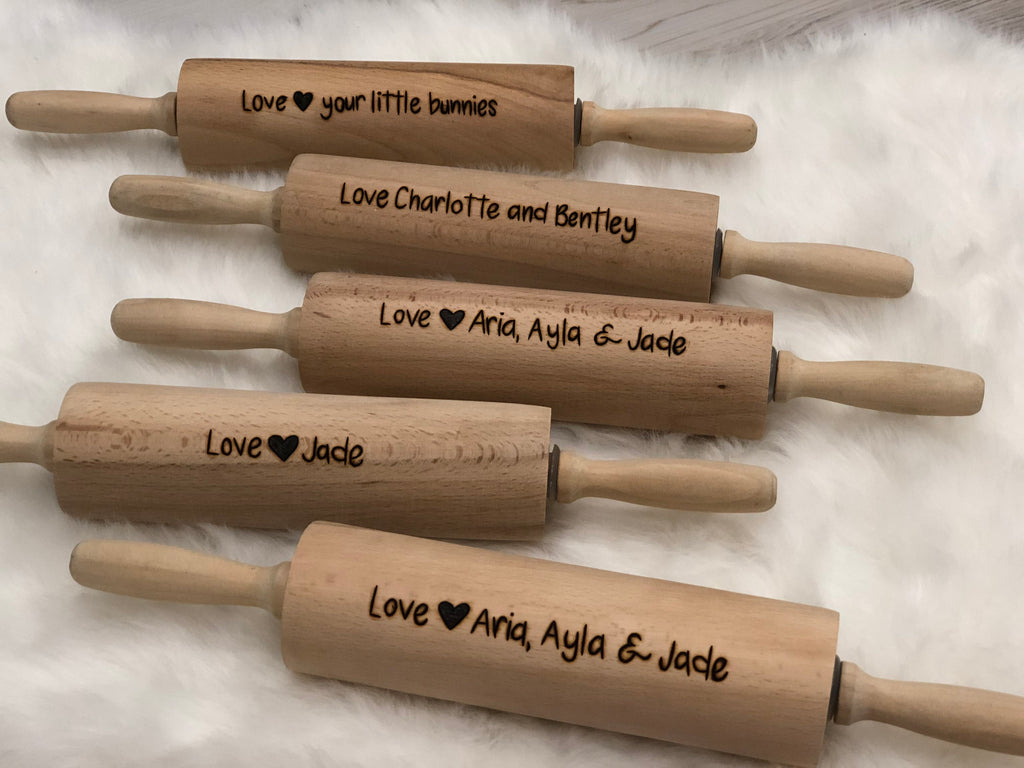Five personalized wooden rolling pins with custom engraved messages resting on white fabric background