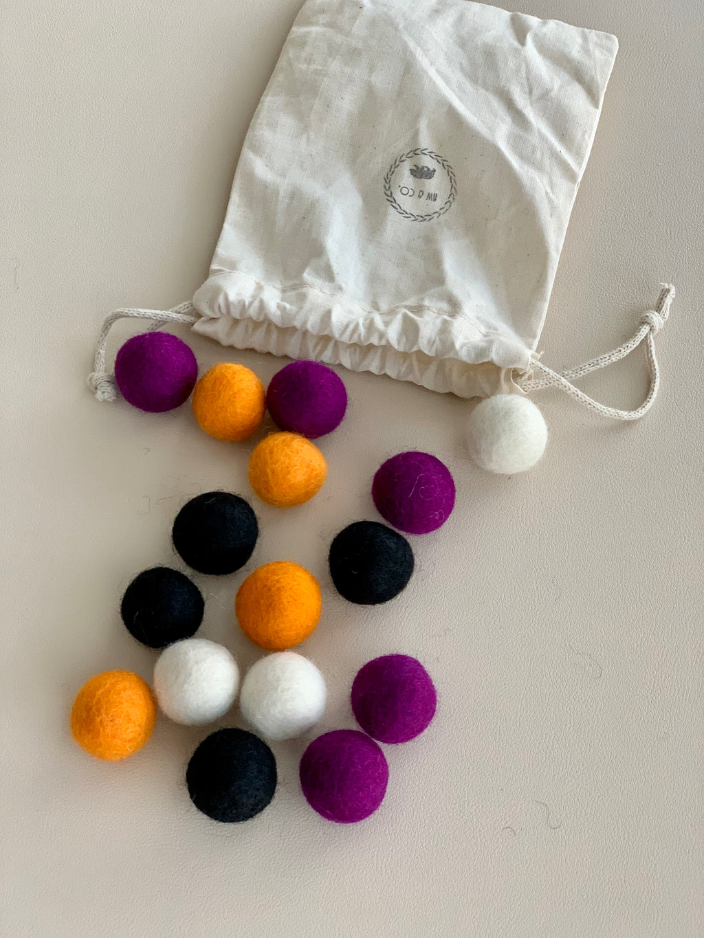 Assorted Halloween-themed felt balls in black, orange, purple, and white with drawstring cotton pouch on cream surface