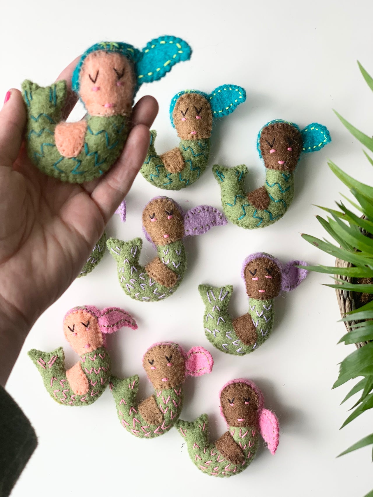 Handmade felt mermaid with turquoise hair and green tail, held in hand with more colorful felt mermaids on white surface
