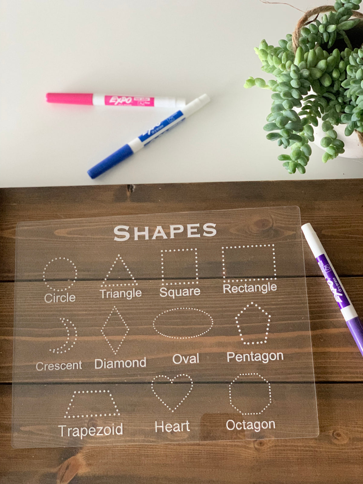Clear acrylic dry erase board with engraved shape outlines and labels for tracing on wood surface with markers and plant nearby