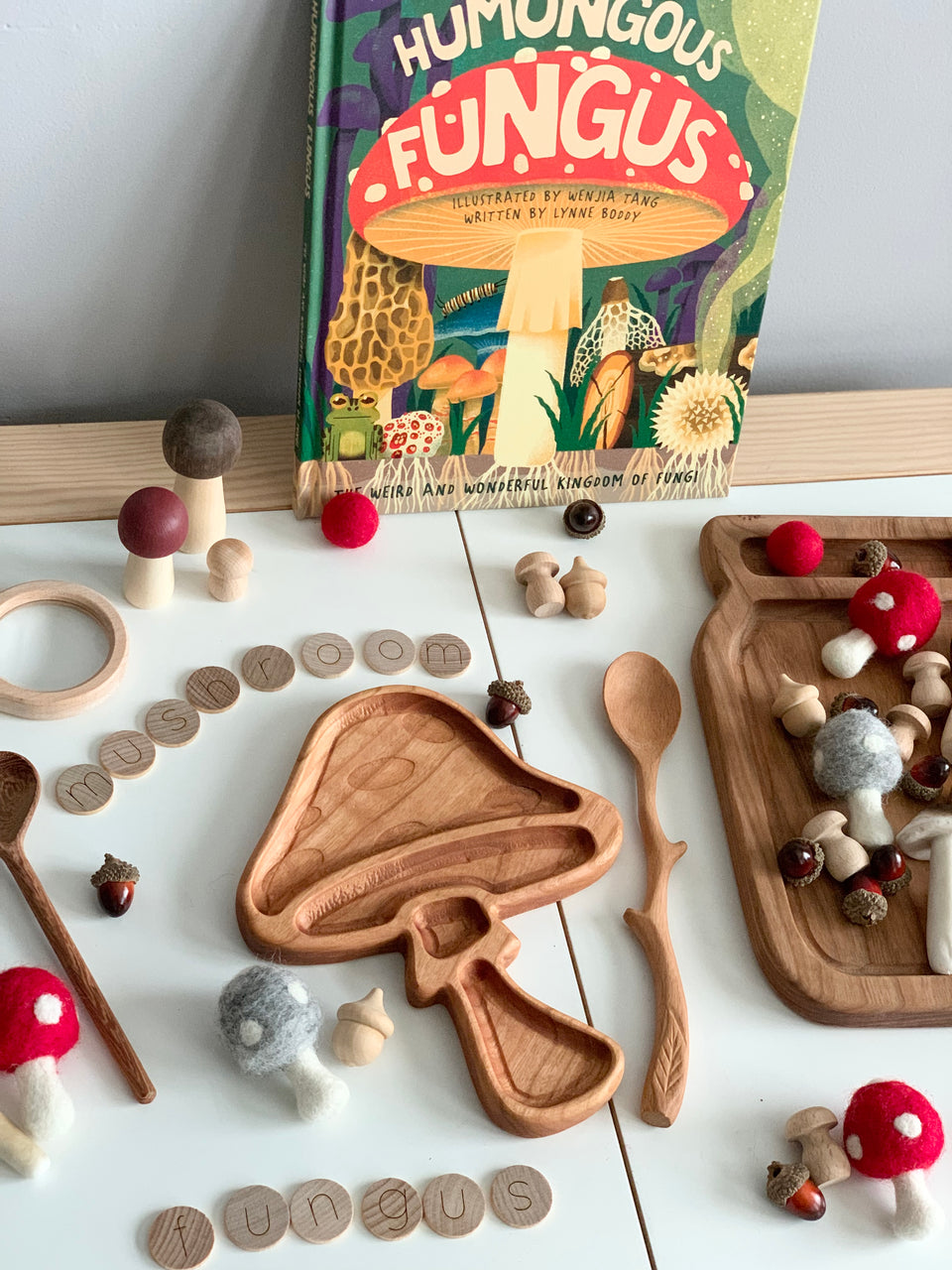 Wooden mushroom-shaped sensory tray with small wooden and felt mushrooms, acorns, and a mushroom-themed book on white surface