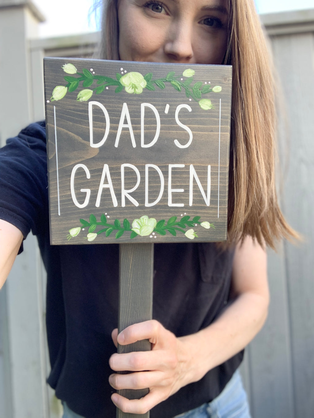 Small square wooden garden sign with white letters reading "DAD'S GARDEN" and floral decorations held by person outdoors