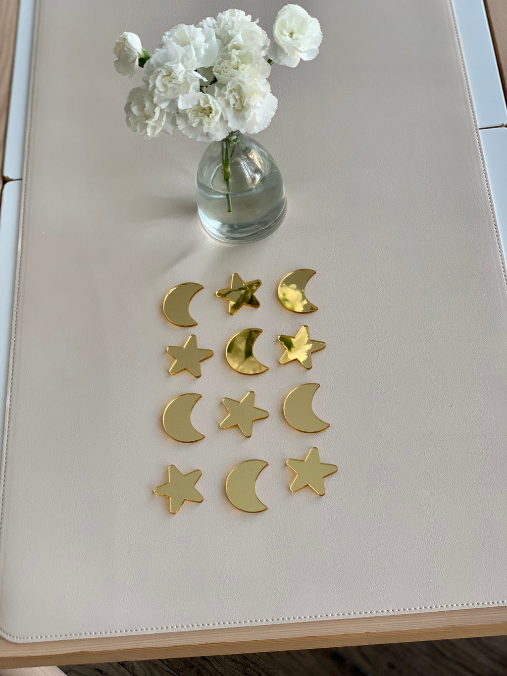 Set of 12 single-sided gold acrylic moons and stars arranged in rows on pale beige surface with small vase of white flowers