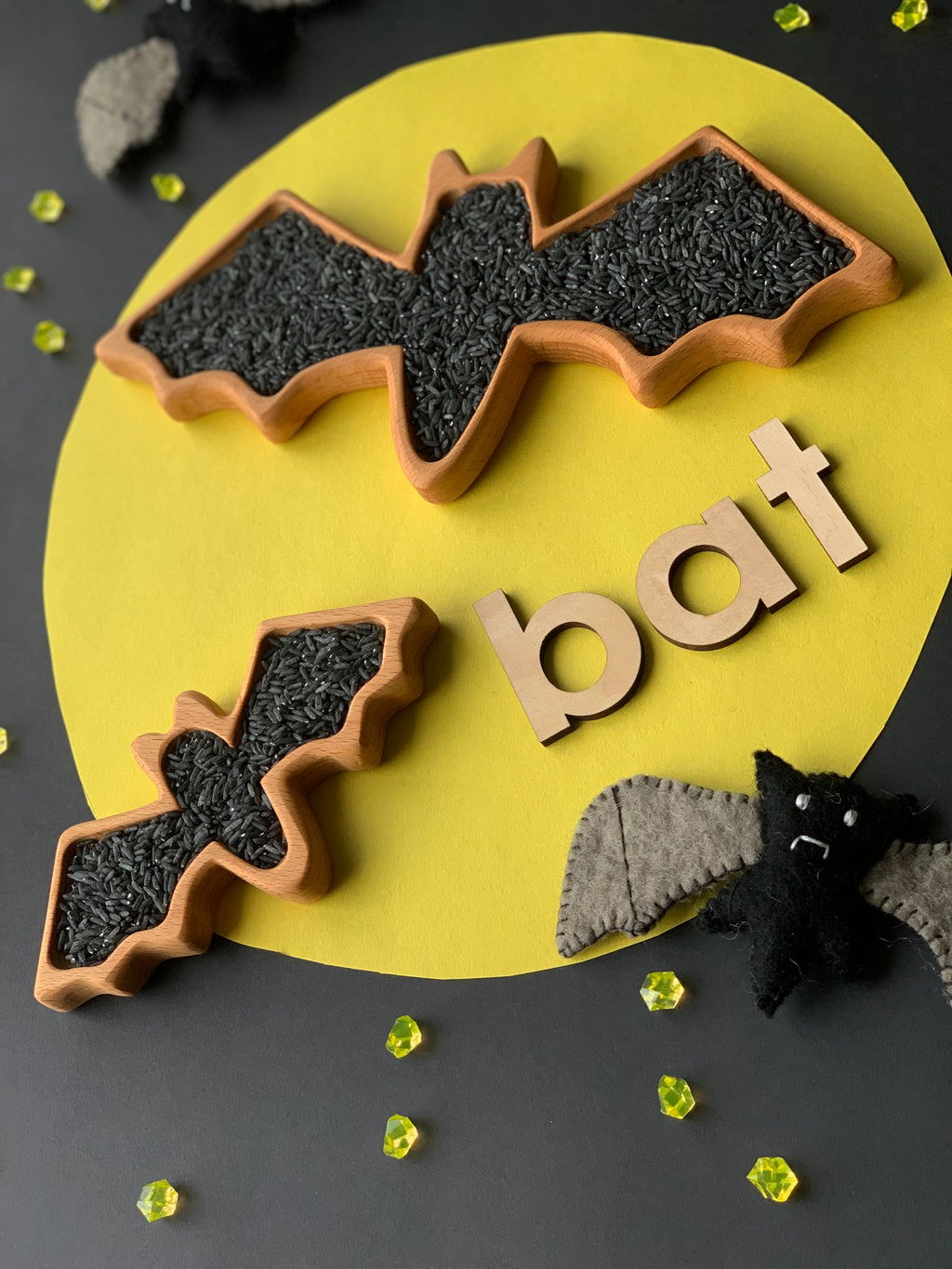 Two wooden bat-shaped sensory trays filled with black rice on yellow and black background with wooden letters spelling bat