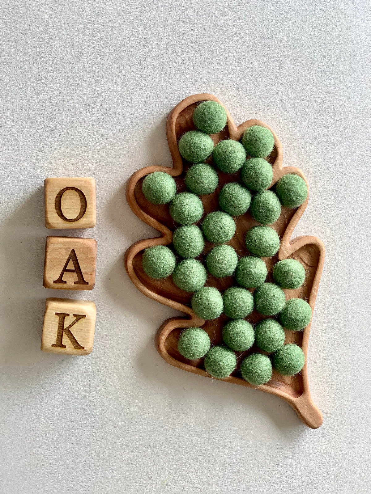 Oak leaf-shaped wooden sensory tray filled with green felt balls next to wooden blocks spelling OAK on white surface