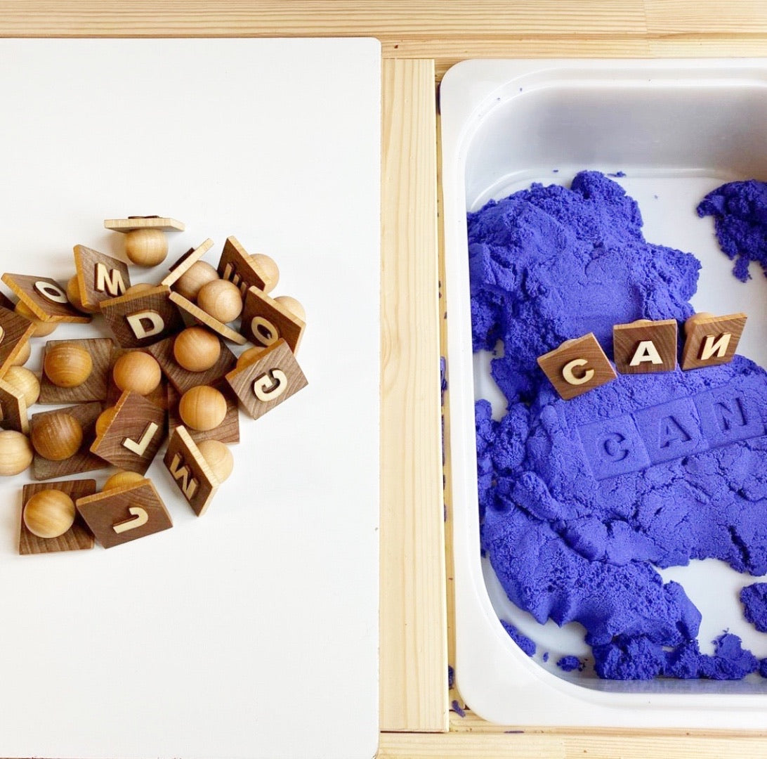 Natural wood alphabet play dough stampers with engraved letters spelling CAN on purple play dough in divided wooden tray