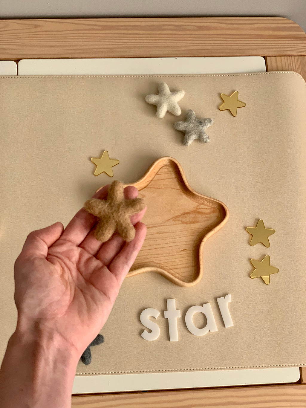 Hand holding small brown felt star above wooden star-shaped tray with gray and white felt stars and gold stars on beige surface