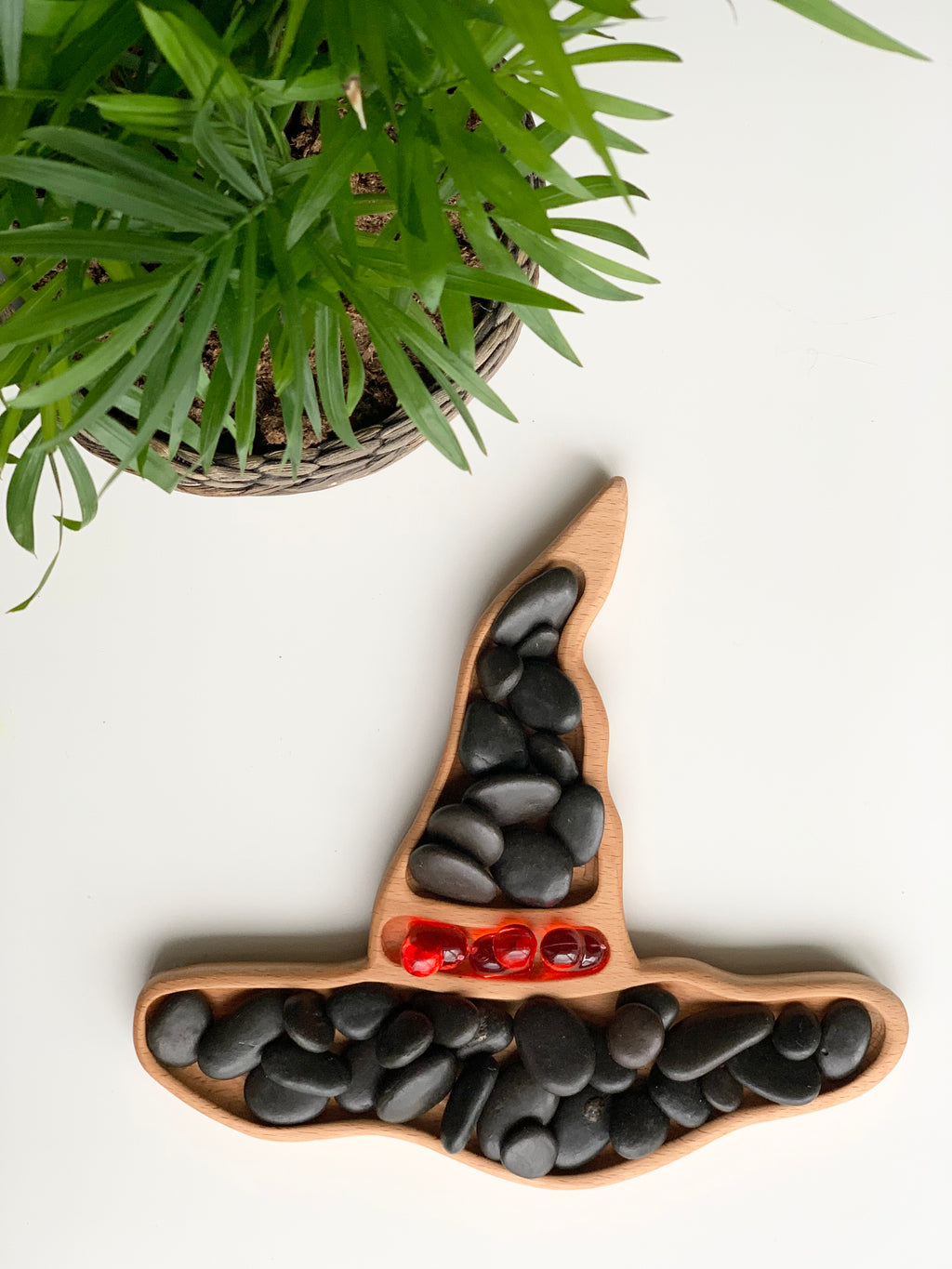 Witch hat shaped wooden sensory tray filled with black and red smooth stones on white surface near green plant