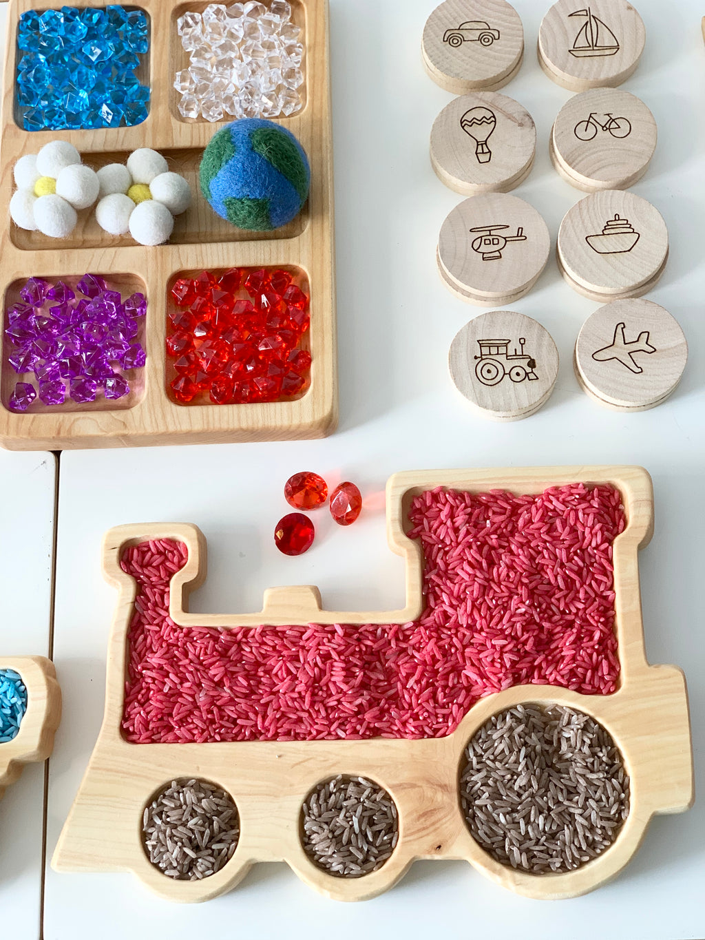 Natural wooden train-shaped sensory tray filled with red and brown rice next to wooden discs with vehicle illustrations