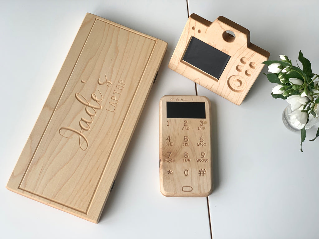 Set of natural wooden toy laptop, camera with engraved buttons and screen, and a phone on white surface with green plant
