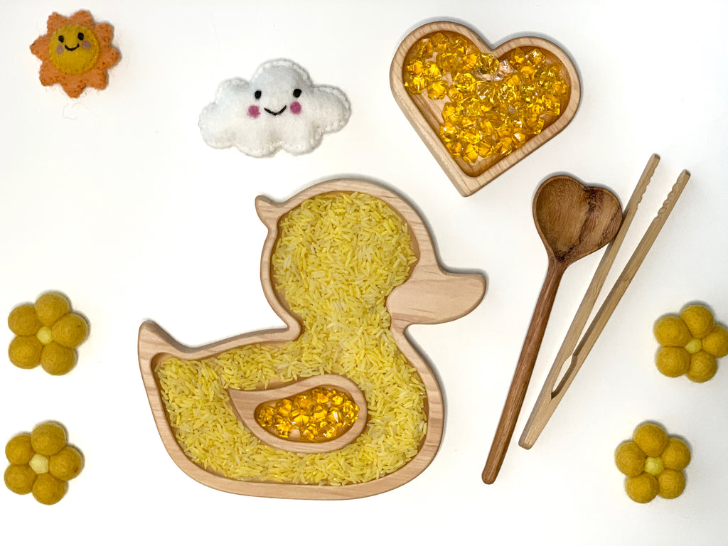 Wooden duck-shaped sensory tray filled with yellow rice and amber beads alongside wooden spoon, tongs, and felt sun and cloud decorations